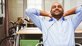 A man in an office, sitting in a chair, leaning back with his hands behind his head, relaxing.
